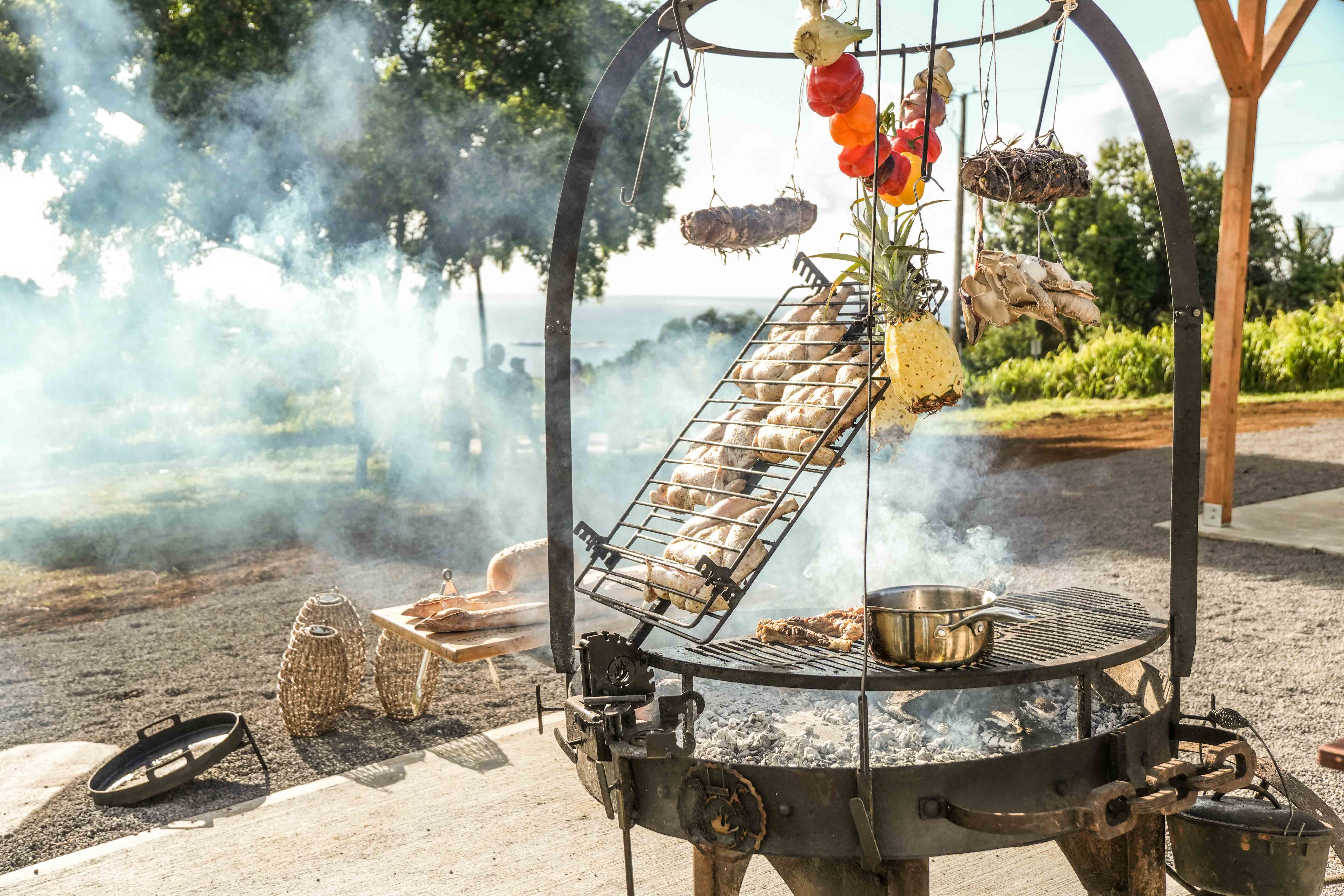 The asado fire wheel with hanging vegetables, smoke rising, and the ocean beyond